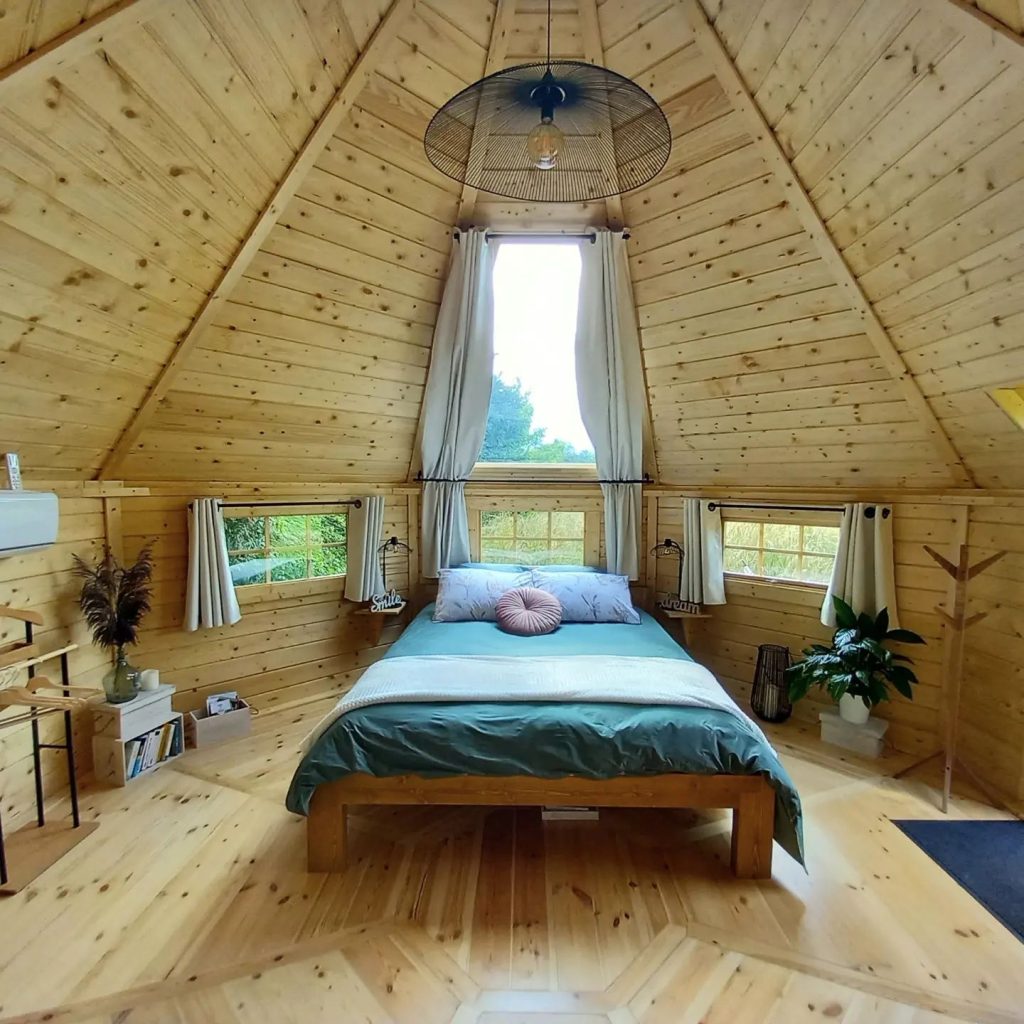Cozy log cabin bedroom with wooden interior, green bed, and natural light through tall window, serene atmosphere