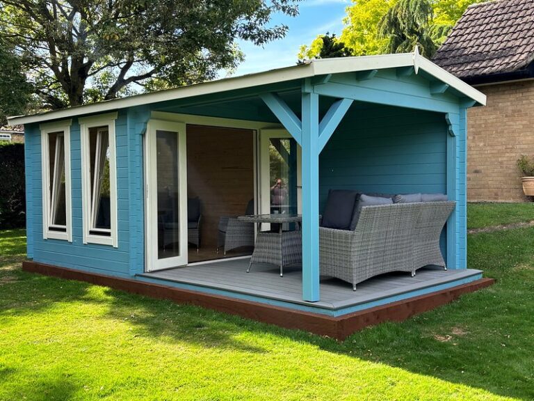 A blue shed with white trim and a covered porch sits on green grass with outdoor furniture inside and a tree and house in the background.