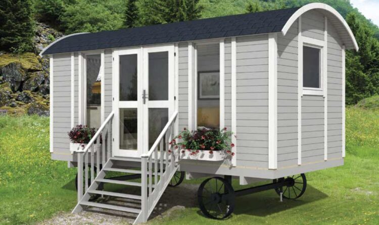 A small grey shepherds hut on wheels with white trim and black roof in a grassy field with flowers and trees