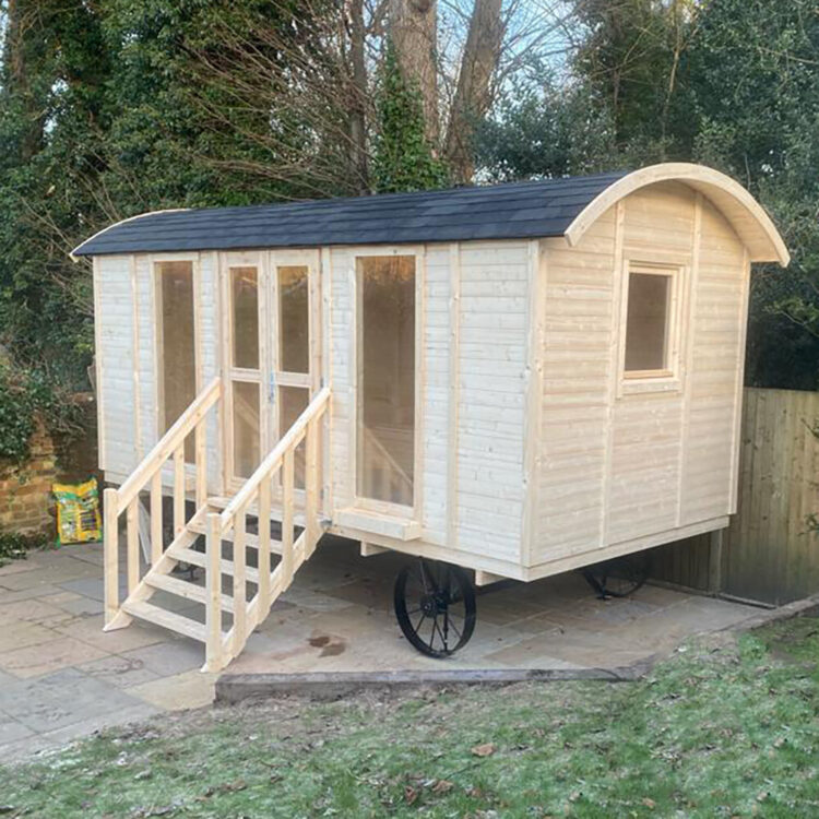 A modern natural shepherds hut with light wood and black roof on wheels in a backyard setting with trees and grass
