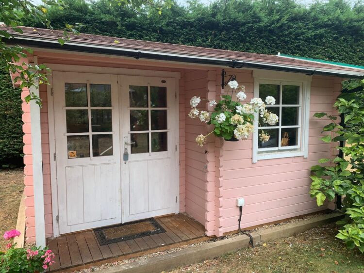A small pink shed with white doors and window, flowers, and wooden porch in a garden setting, resembling a shepherds hut or cabin
